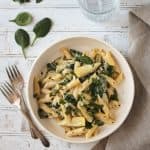 Bowl of vegan spinach artichoke pasta in a white bowl next to two forks.