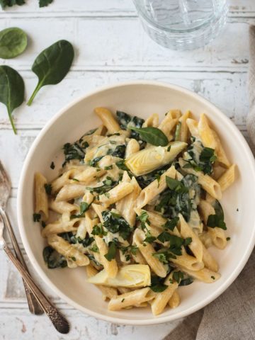 Bowl of vegan spinach artichoke pasta in a white bowl next to two forks.