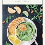 Pumpkin seed pesto in a bowl on a plate with crackers and two lemon halves.