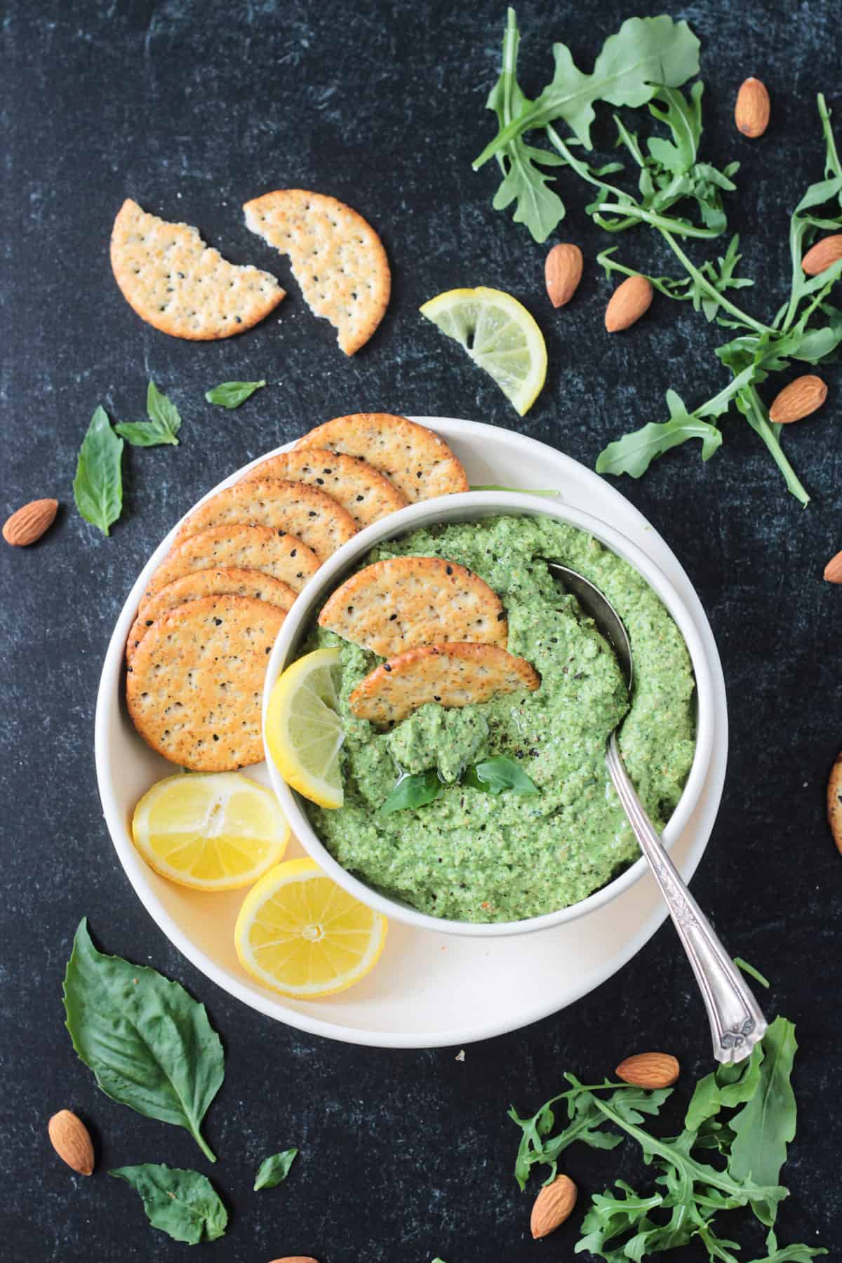 Pumpkin seed pesto in a bowl on a plate with crackers and two lemon halves.