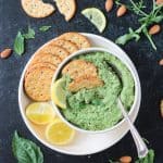 Pumpkin seed pesto in a bowl on a plate with crackers and two lemon halves.