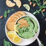 Pumpkin seed pesto in a bowl on a plate with crackers and two lemon halves.