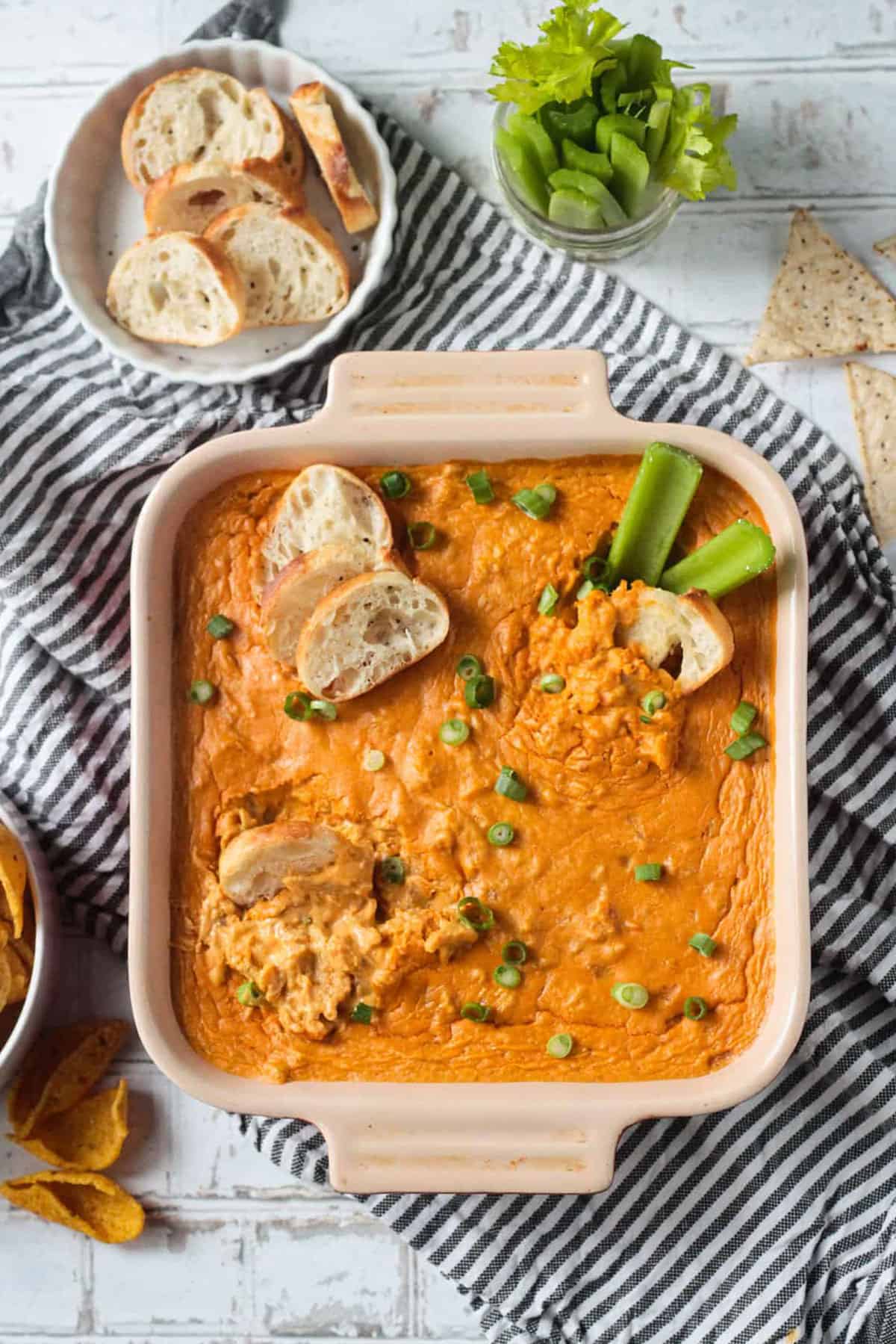 Baguette slices dipped in Vegan Buffalo Chicken Dip in a square baking dish topped with green onions.