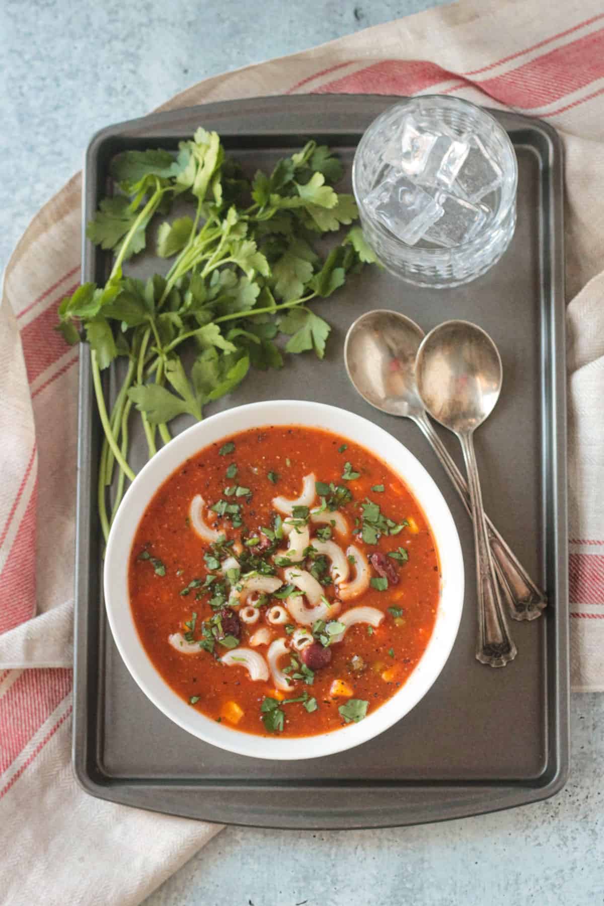 Bowl of soup on a tray with two spoons, glass of water, and bunch of fresh parsley.