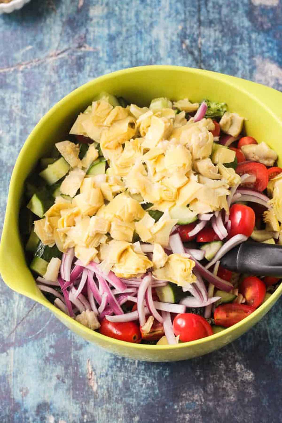 Chopped artichokes, tomatoes, cucumbers, and sliced red onion in a green mixing bowl.