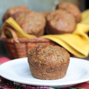 One muffin on a white plate in front of a basket of vegan zucchini muffins.