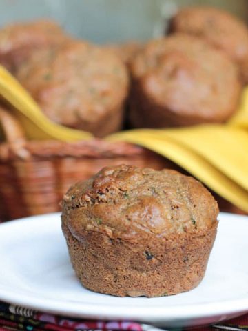 One muffin on a white plate in front of a basket of vegan zucchini muffins.