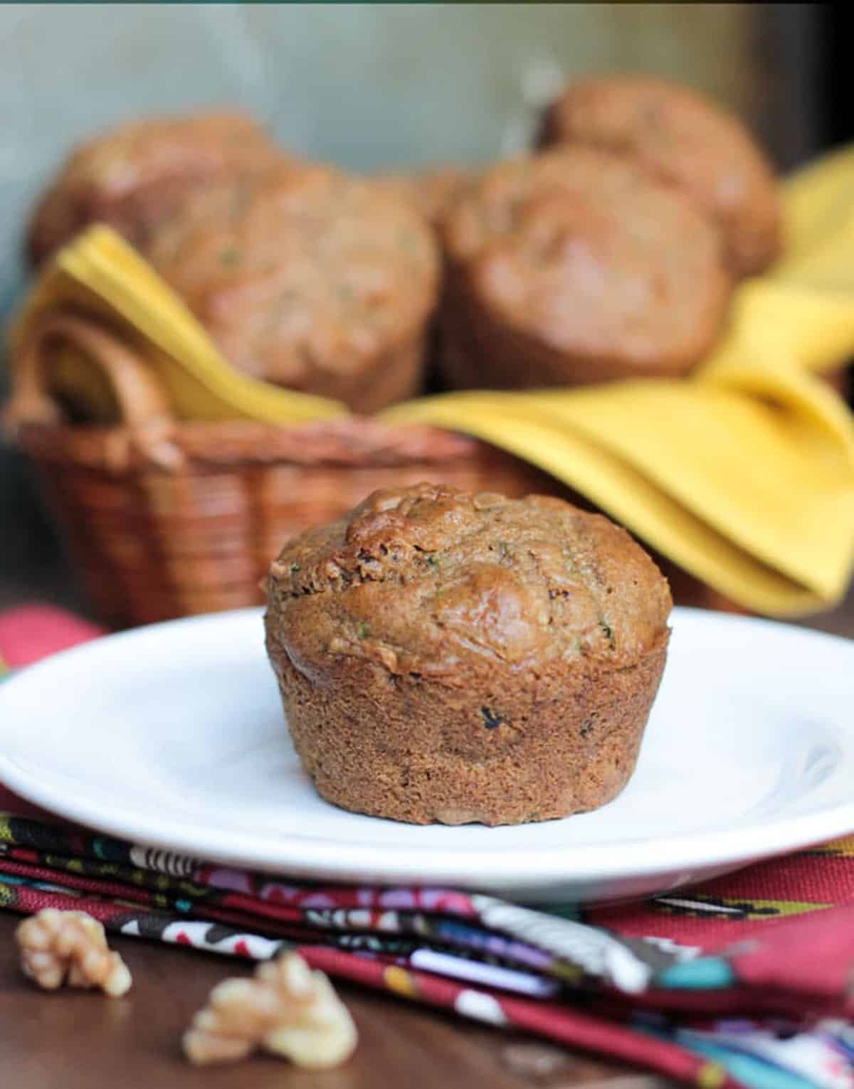 One muffin on a white plate in front of a basket of vegan zucchini muffins.