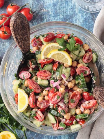 Two wooden serving spoons in a bowl of cucumber chickpea salad.