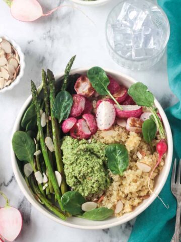 Quinoa veggie bowl with asparagus, radishes, fresh spinach, and pesto in a white flat bowl.