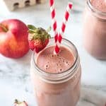 One pink smoothie in a glass next to an apple and halved strawberries.