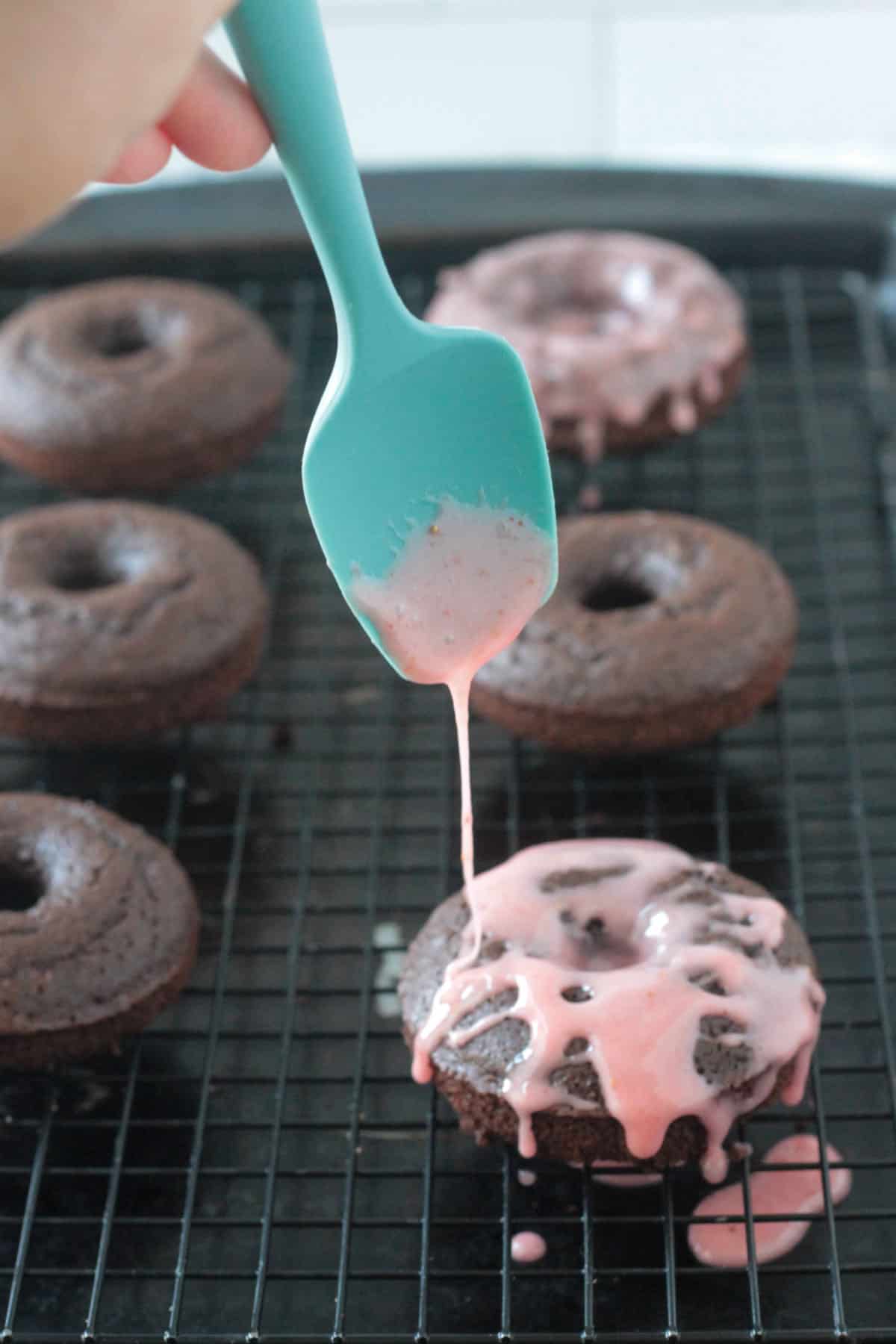 Pink icing being drizzled onto a chocolate donut from a small blue spatula.
