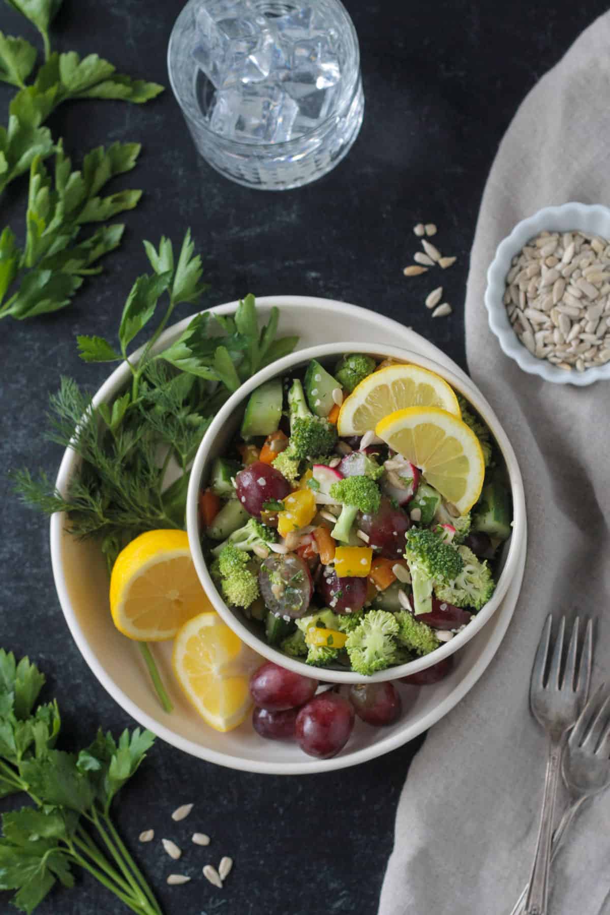 Bowl of salad on a plate with fresh herbs, lemon wedges, and a bunch of red grapes.