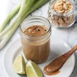 Peanut sauce in a glass jar on a white plate with lime wedges.