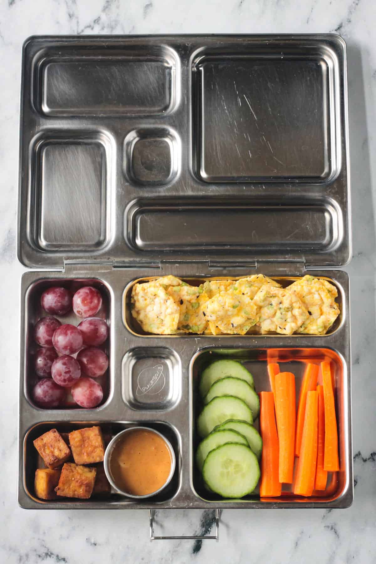 5 compartment lunchbox with carrots, cucumber, baked tofu, peanut sauce, red grapes, popcorn chips.