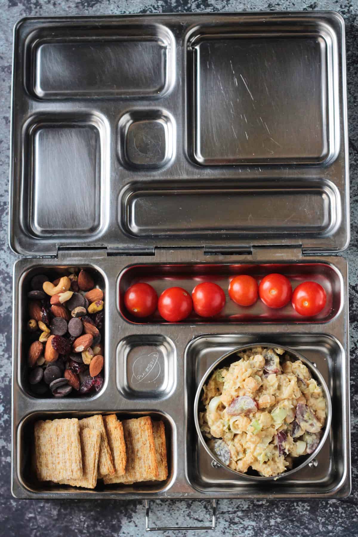 Metal lunchbox with chickpea salad, wheat crackers, trail mix, and grape tomatoes.