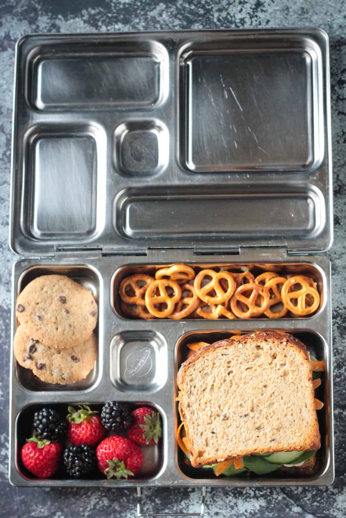 Vegan lunch box with hummus veggie sandwich, strawberries, blackberries, pretzels, chocolate chip cookies.