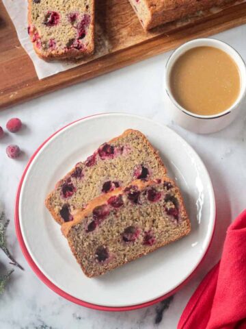 Two slices of vegan cranberry bread on a white plate next to a cup of coffee.