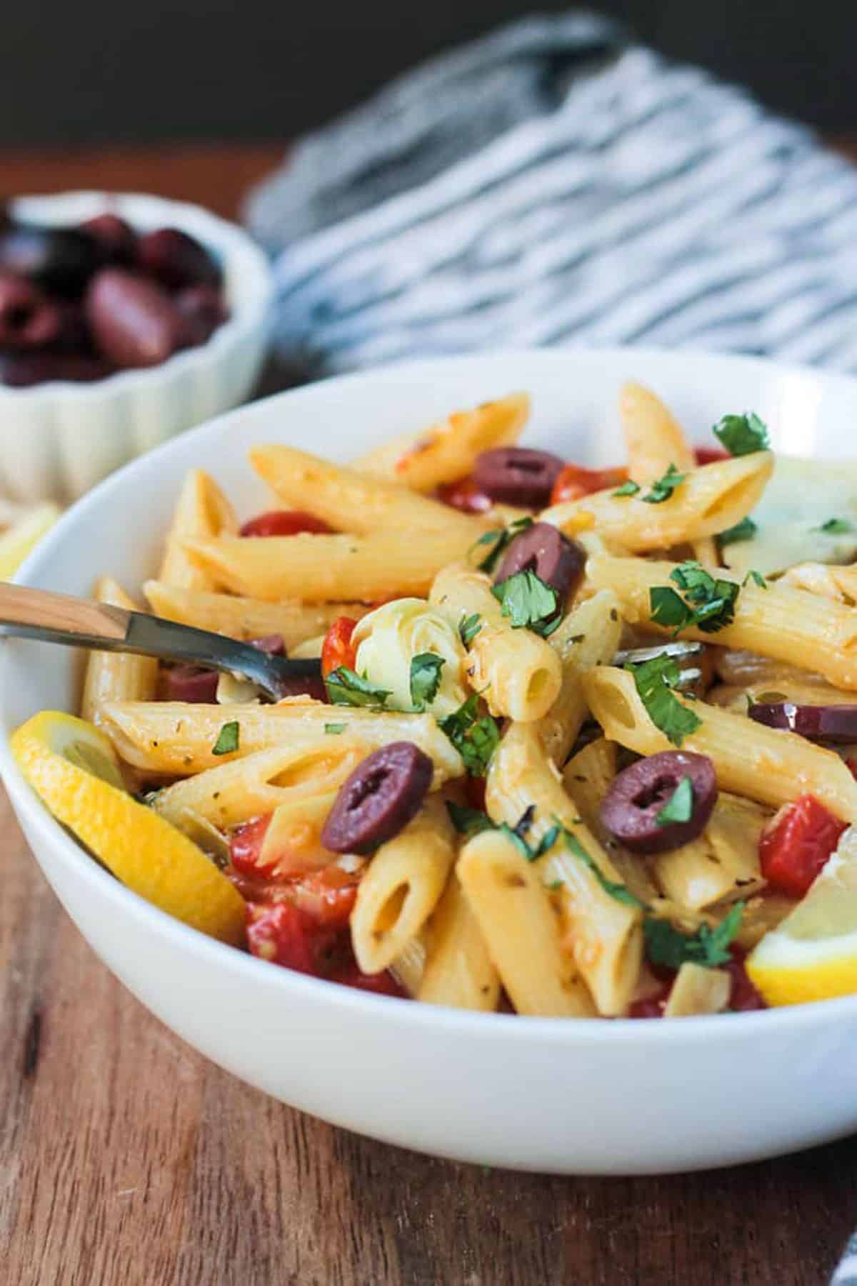 Penne pasta with artichokes, red peppers, and olives in a white bowl.