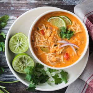 Red Curry Thai Noodle Soup in a white bowl next to two lime halves and cilantro leaves.