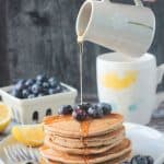 Syrup being poured onto a stack of 5 oat flour pancakes topped with blueberries.