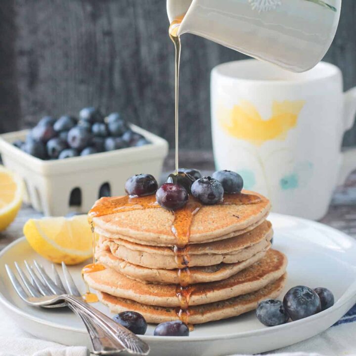Syrup being poured onto a stack of 5 oat flour pancakes topped with blueberries.