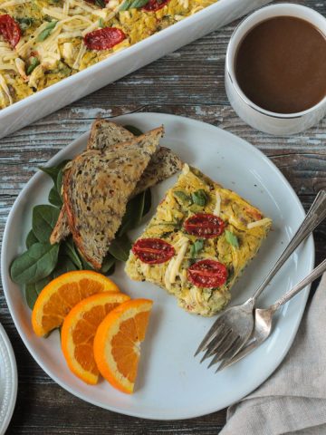Slice of vegan breakfast casserole on a plate with toast, orange slices, and two forks.