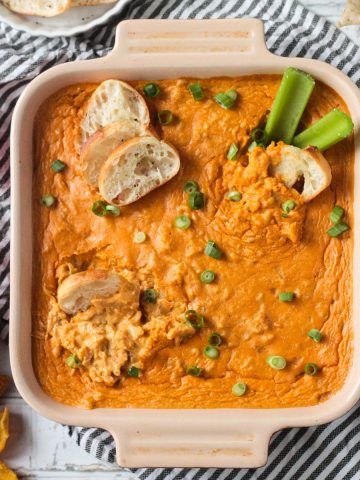Baguette slices dipped in Vegan Buffalo Chicken Dip in a square baking dish topped with green onions.