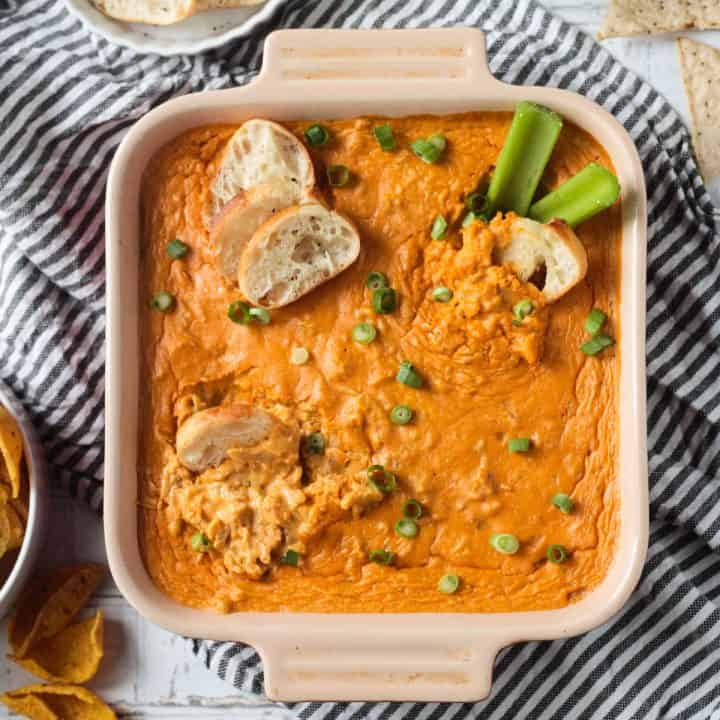 Baguette slices dipped in Vegan Buffalo Chicken Dip in a square baking dish topped with green onions.