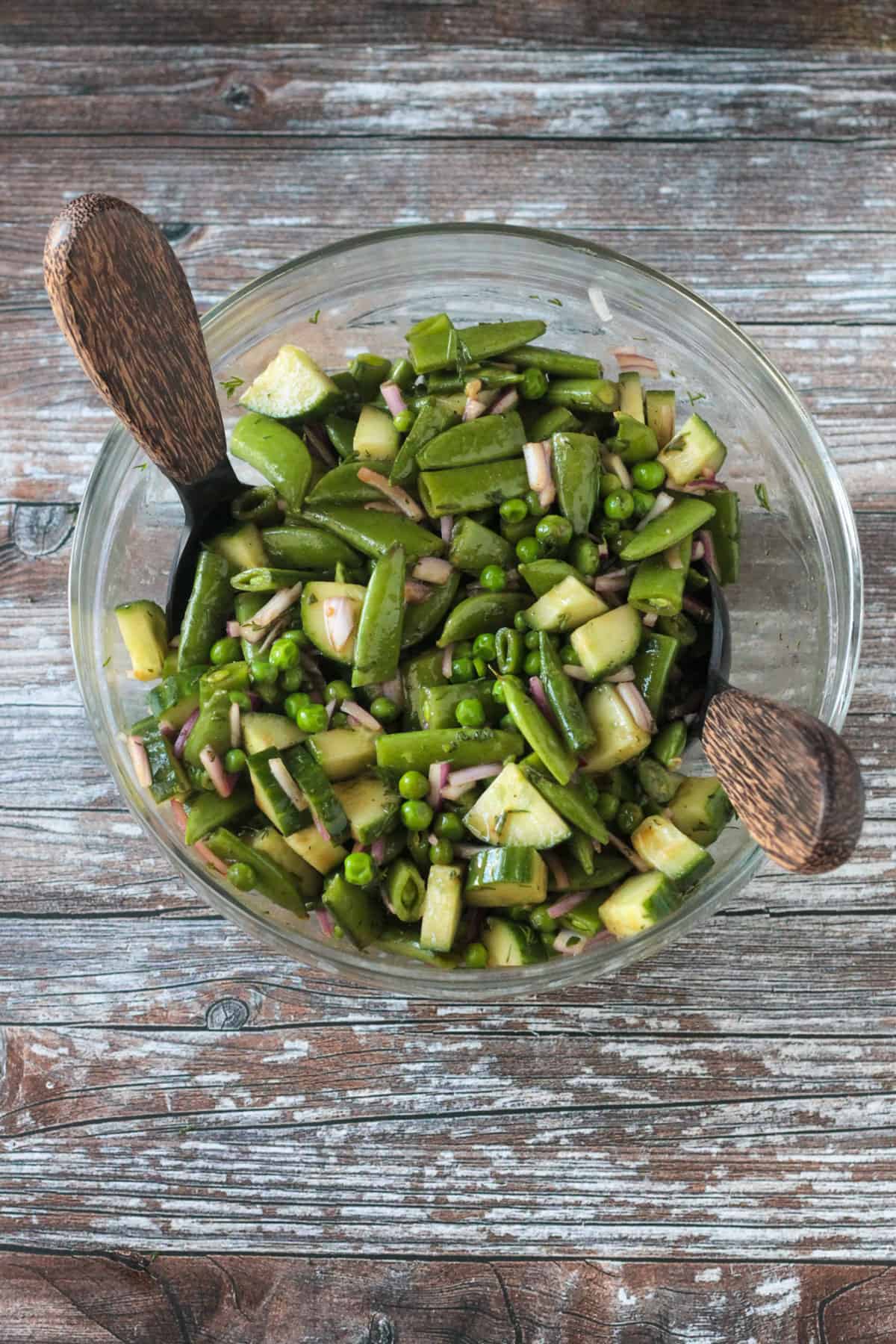Snap peas, sweet peas, cucumbers, onion, fresh herbs, and dressing mixed in a glass bowl.