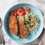 Two breaded vegan chicken cutlets on a blue plate with rice, peas, and tomatoes.