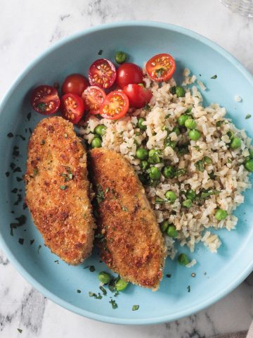 Two breaded vegan chicken cutlets on a blue plate with rice, peas, and tomatoes.