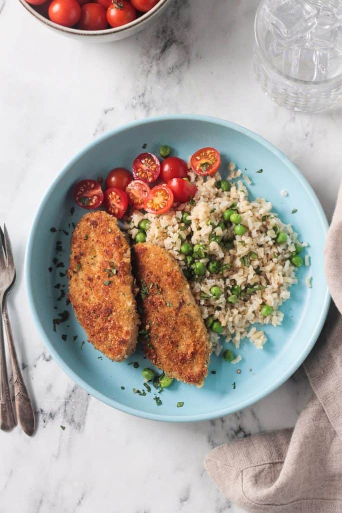 Two breaded vegan chicken cutlets on a blue plate with rice, peas, and tomatoes.