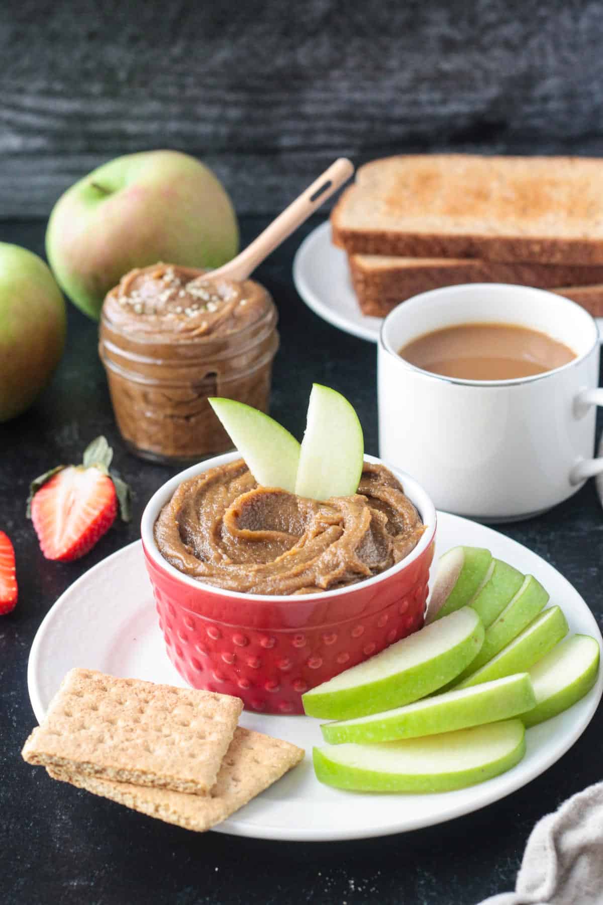 Two apple slices in a bowl of cookie butter next to a cup of coffee and stack of toast.