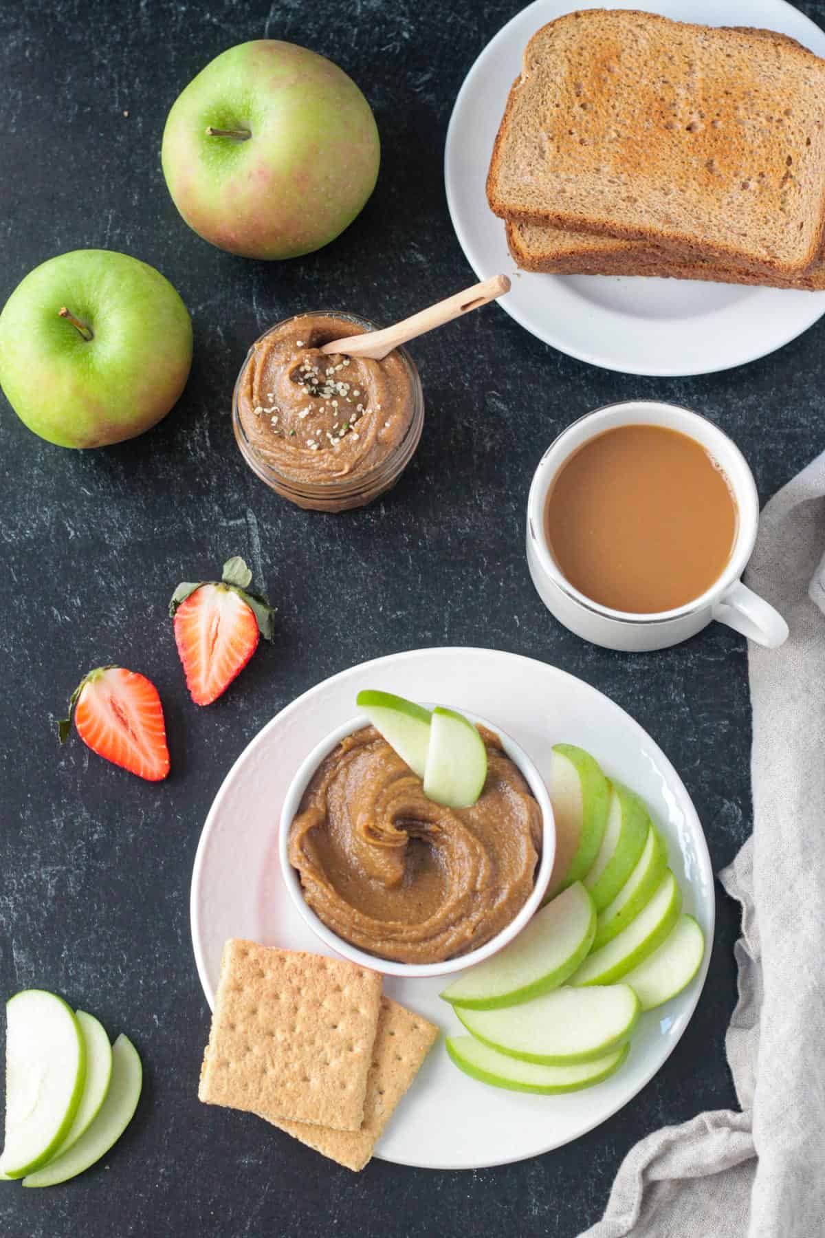 Cookie butter in a bowl on a plate with apples and crackers next to coffee and toast.