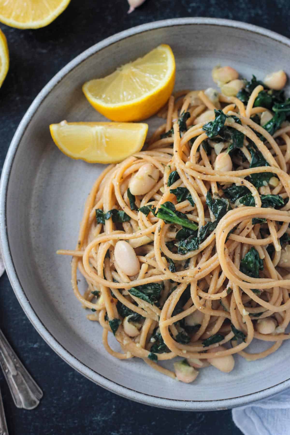 Close up of white beans and kale mixed with spaghetti noodles on a gray plate.