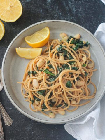 Spaghetti past with white beans kale on a gray plate with two lemon wedges.