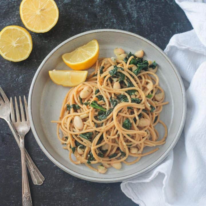 Spaghetti past with white beans kale on a gray plate with two lemon wedges.