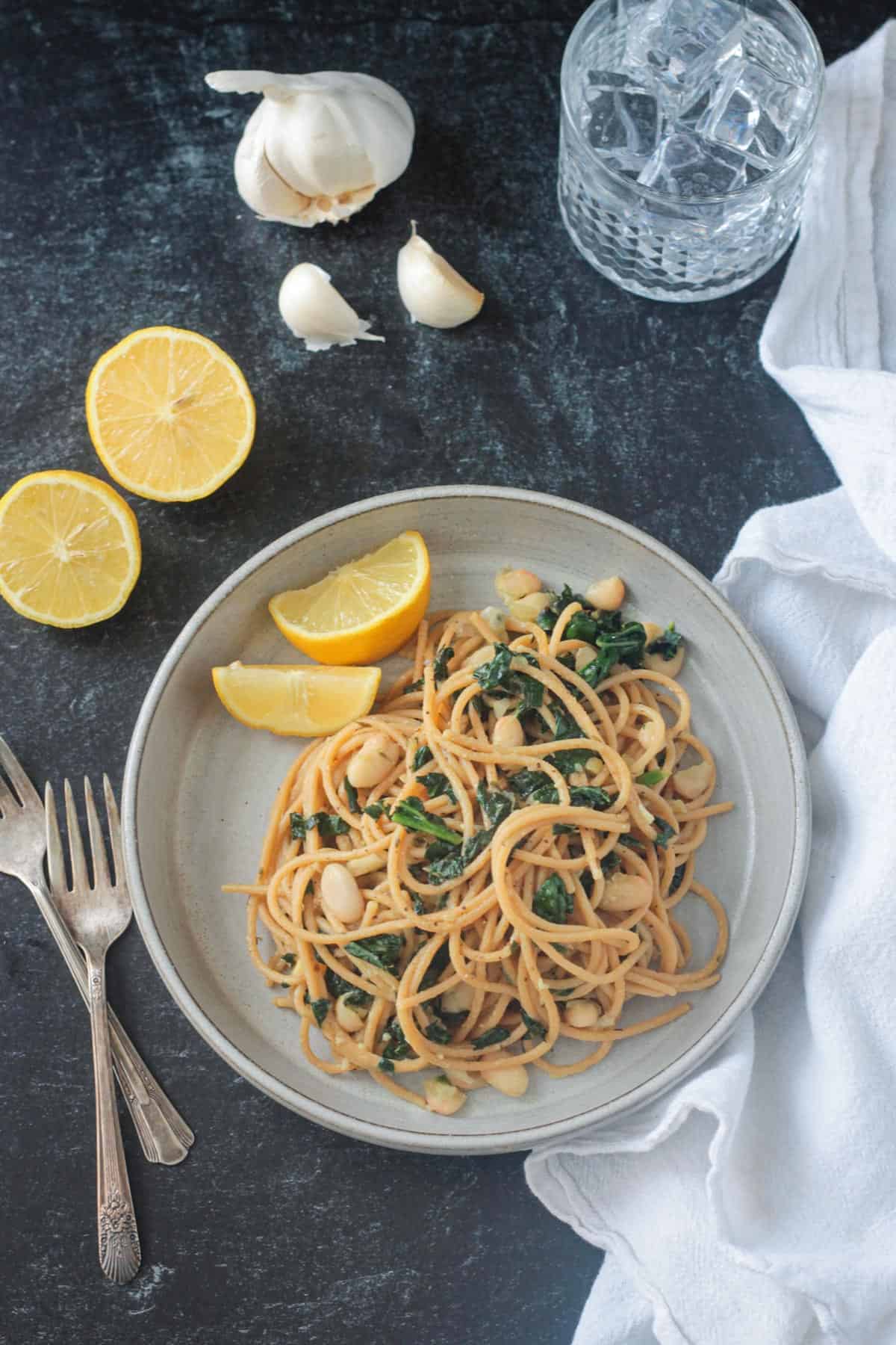 Spaghetti past with white beans kale on a gray plate with two lemon wedges.