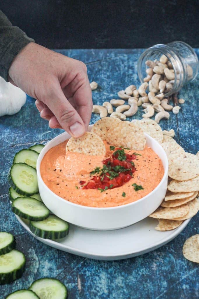 Adult hand dipping a tortilla chip into bowl of red pepper spread.