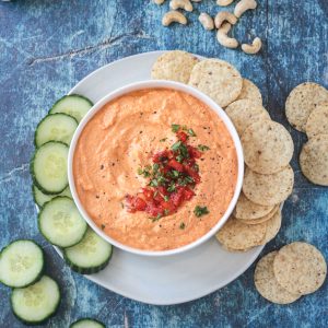 Bowl of roasted red pepper cashew dip on a plate with sliced cucumbers and tortilla chips.