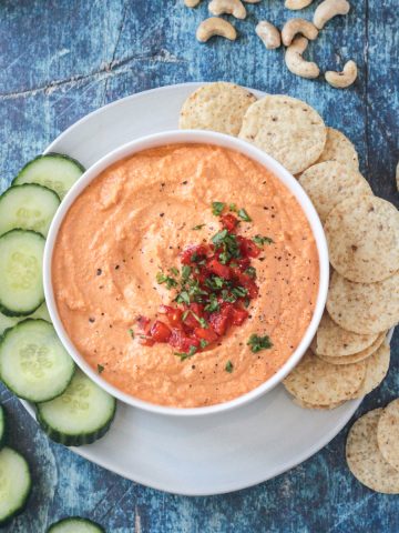 Bowl of roasted red pepper cashew dip on a plate with sliced cucumbers and tortilla chips.