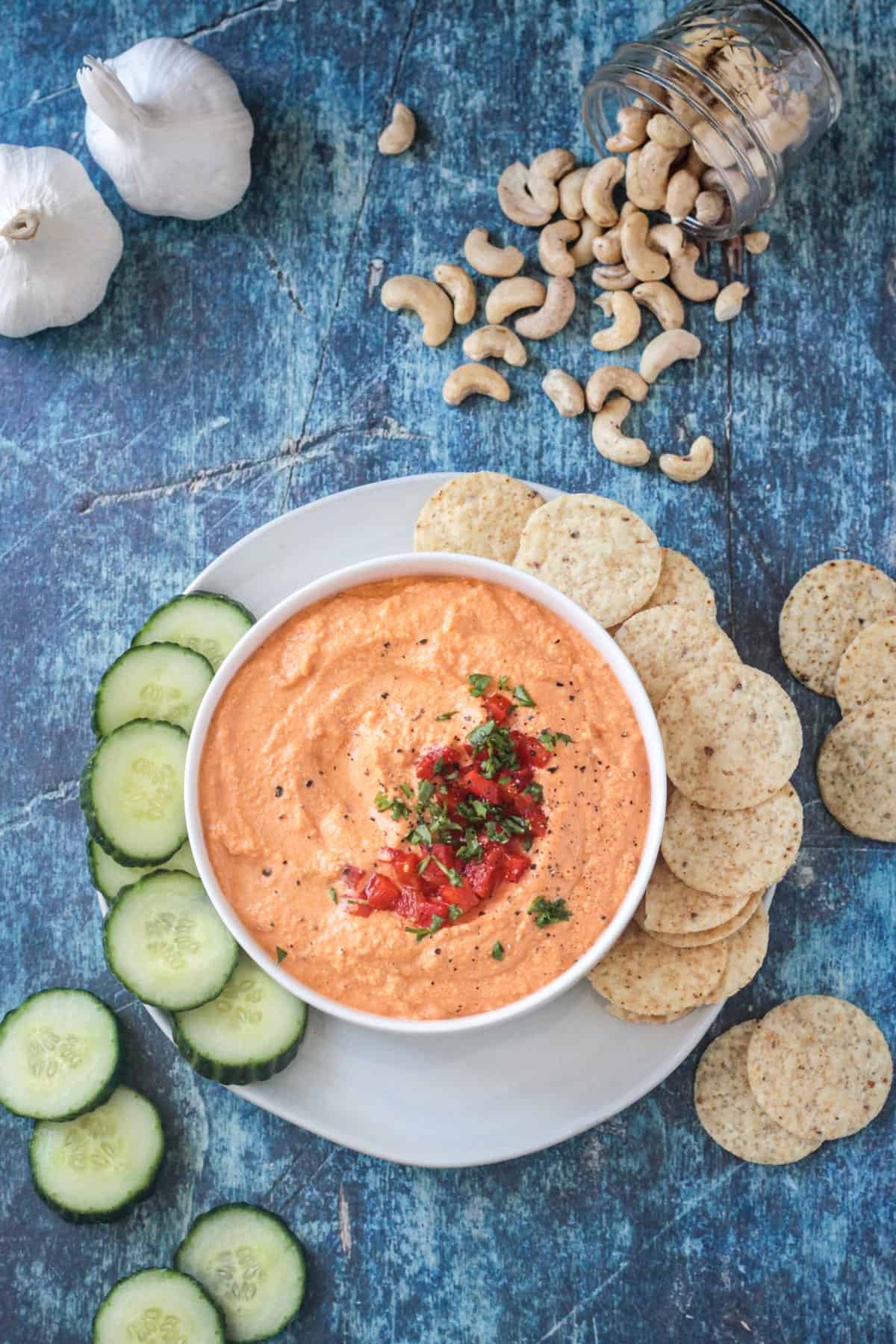 Bowl of roasted red pepper cashew dip on a plate with sliced cucumbers and tortilla chips.