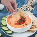 Adult hand dipping a tortilla chip into bowl of red pepper spread.