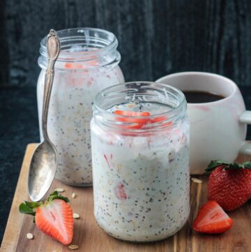 Two jars of strawberry overnight oats with coconut milk in front of a cup of coffee.