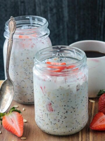 Two jars of strawberry overnight oats with coconut milk in front of a cup of coffee.
