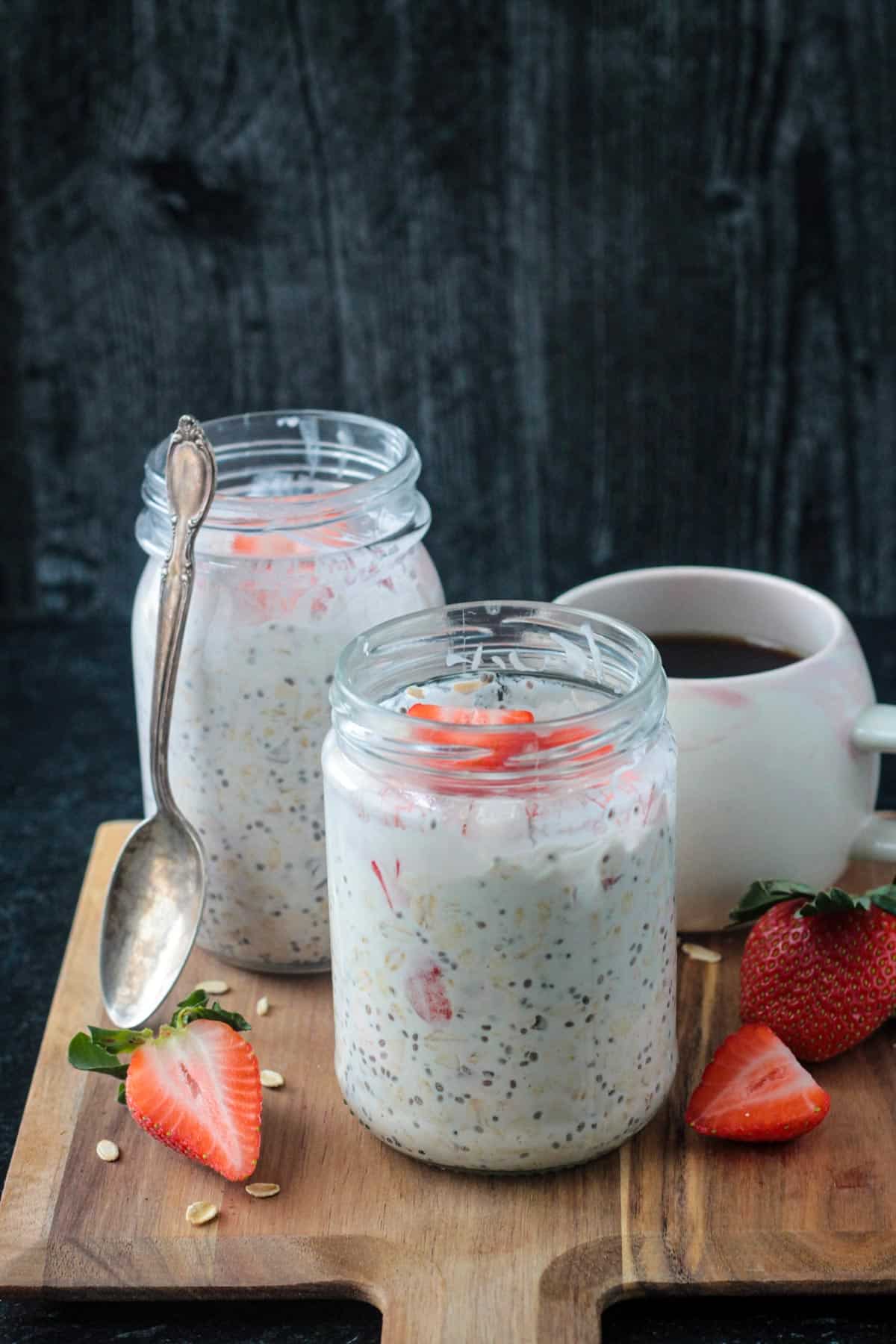 Two jars of strawberry overnight oats with coconut milk in front of a cup of coffee.