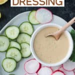Tahini goddess dressing in a small white bowl surrounded by sliced cucumbers and radishes on a plate.