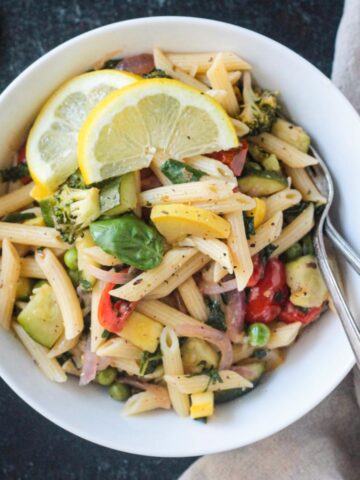 Vegan pasta primavera in a white bowl garnished with two lemon wedges.