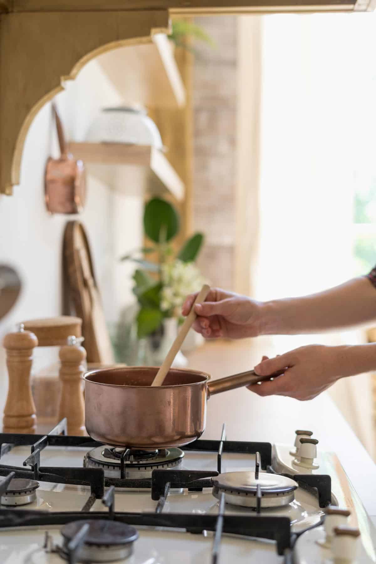Adult hands holding the handle of a pot on the stove while stirring with a wooden spoon.